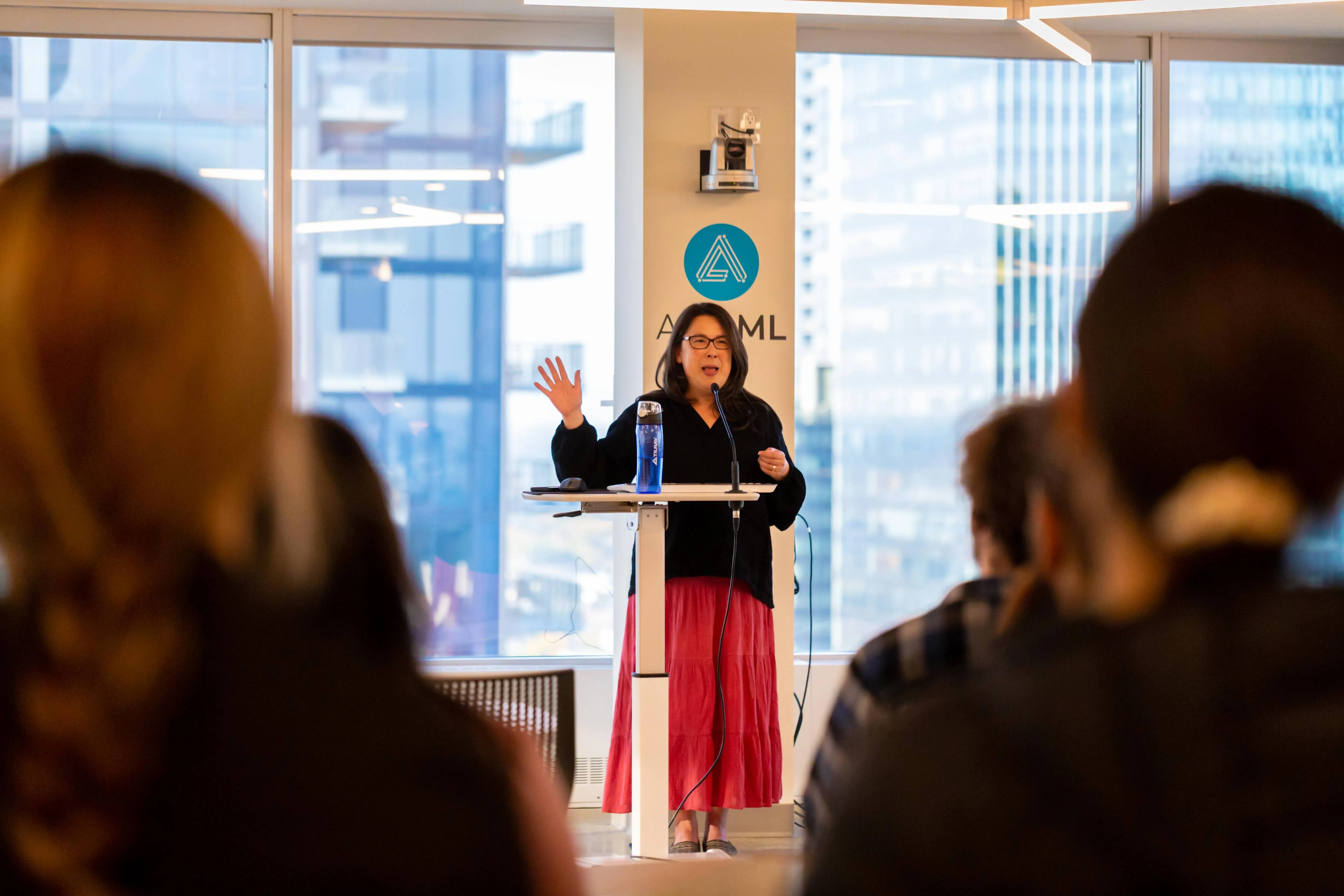 A woman standing at a podium, talking into a microphone with her right hand waving.